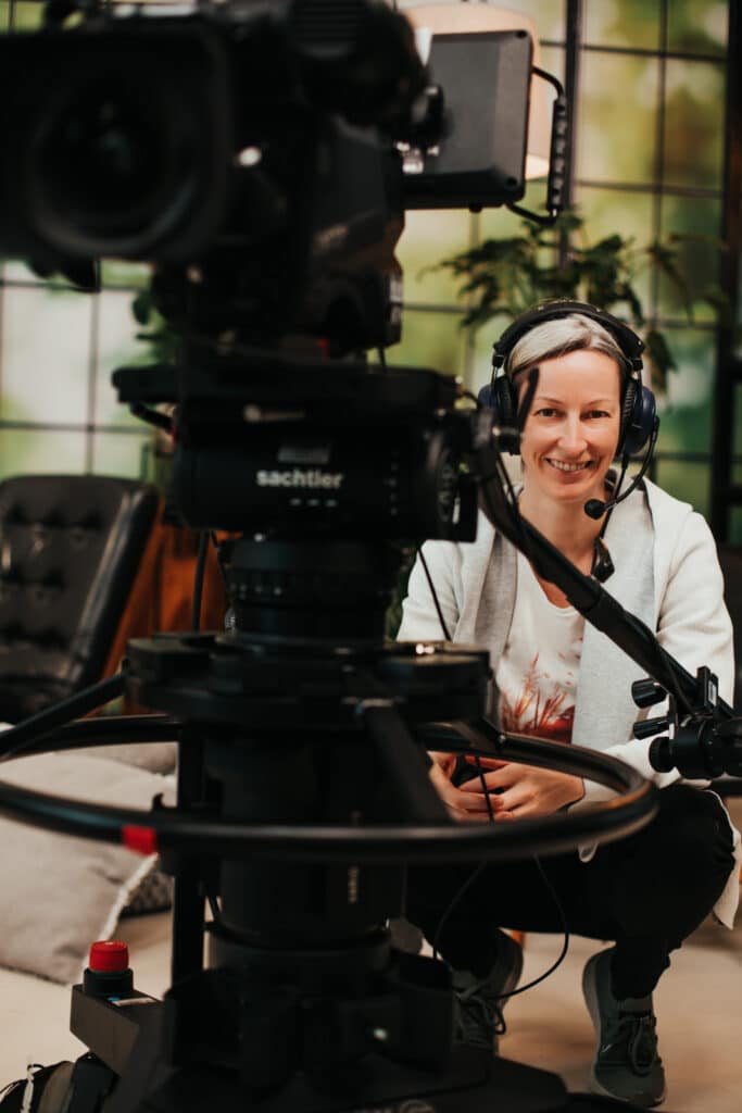 Kamerafrau an einer Kamerapumpe während einer Liveproduktion in der Hocke und lacht in die Kamera